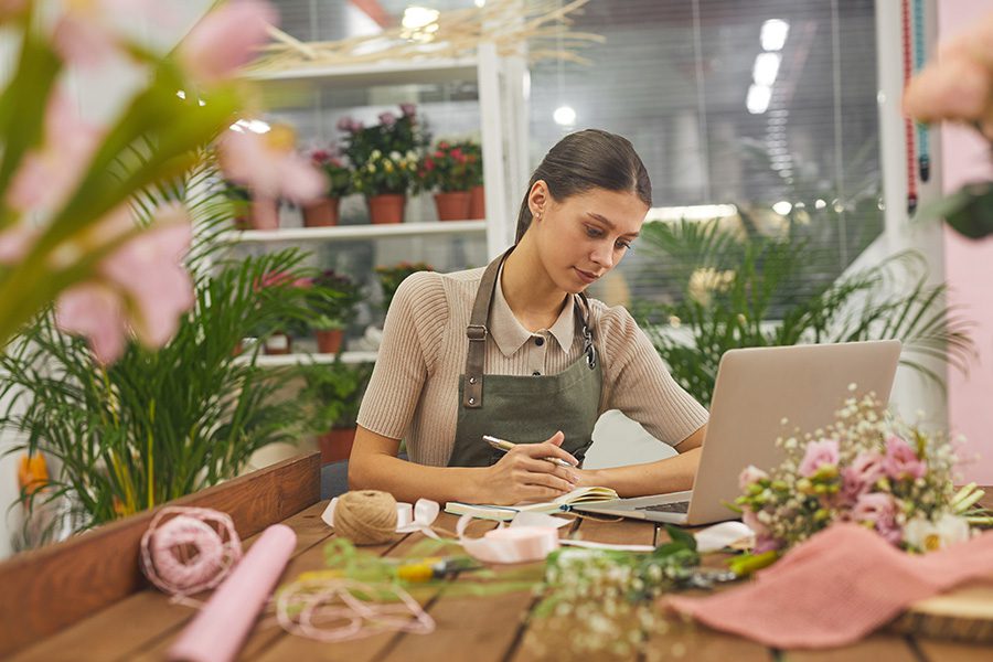 Business Insurance - Female Small Business Owner Managing Accounting Books While Using Laptop at Table in Flower Shop